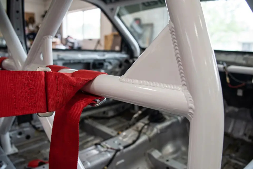 Close-up of a white roll cage weld with a gusset plate, a red harness strap running past it.